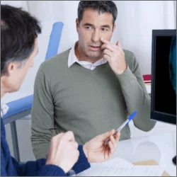Pacjent chory na zapalenie zatok rozmawia z lekarzem w jego gabinecie, na ekranie monitora widać prześwietloną czaszkę z widocznymi drogami oddechowymi