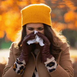 Kobieta ubrana w żółtą czapkę i płaszcz spaceruje w parku i wydmuchuje nos w chusteczkę, cierpi na zapalenie zatok
