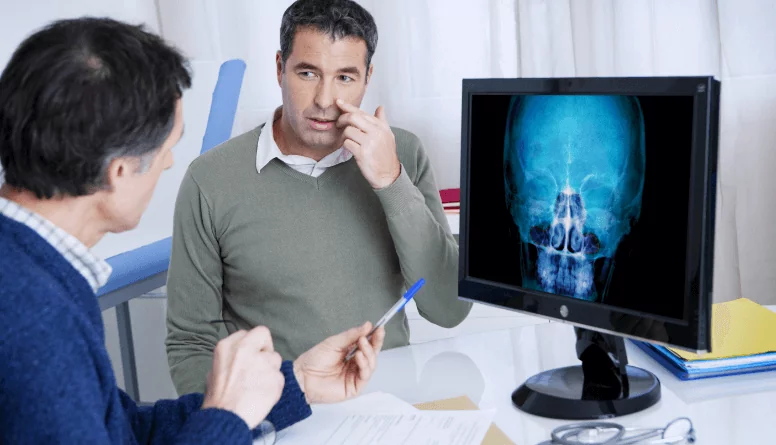 Lekarz konsultuje z pacjentem zalecenia dotyczące postępowania w trakcie zapalenia zatok, na ekranie monitora widać prześwietloną czaszkę z widocznymi drogami oddechowymi